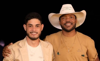 A imagem mostra Júnior e Zé Rodrigo sorrindo e posando lado a lado. O da esquerda tem barba curta, cabelo escuro e veste camiseta clara com um blazer bege. O da direita usa um chapéu branco, camiseta bege e um colar com crucifixo, com o braço apoiado sobre o ombro do outro. Ambos transmitem simpatia e proximidade.